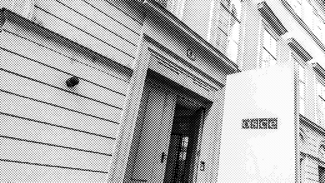 This image shows the entrance to the Organization for Security and Co-operation in Europe (OSCE). The building's facade is light-colored with horizontal grooves and rectangular windows that are symmetrically aligned. Above the entrance, the building's street number ("6") is visible on a small plaque. A white panel with the OSCE logo displayed is positioned near the doorway. The door is slightly open, offering a glimpse into the building's interior, which has glass doors inside.