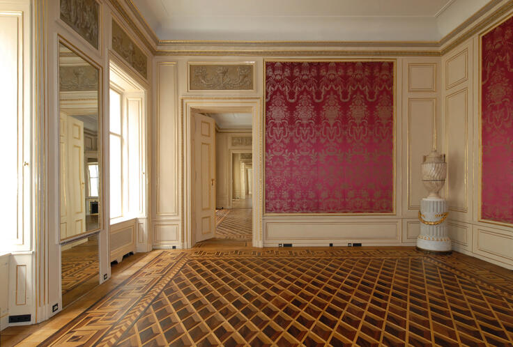 The image shows an elegant, historical interior. The room features intricate parquet flooring with a geometric pattern, ornate wall paneling in cream and gold, and rich red damask wallpaper. Large mirrors and windows reflect light, enhancing the sense of space. A decorative urn on a pedestal adds a classical touch. The open doorways lead to similarly adorned rooms, creating depth and continuity in the architectural design.
