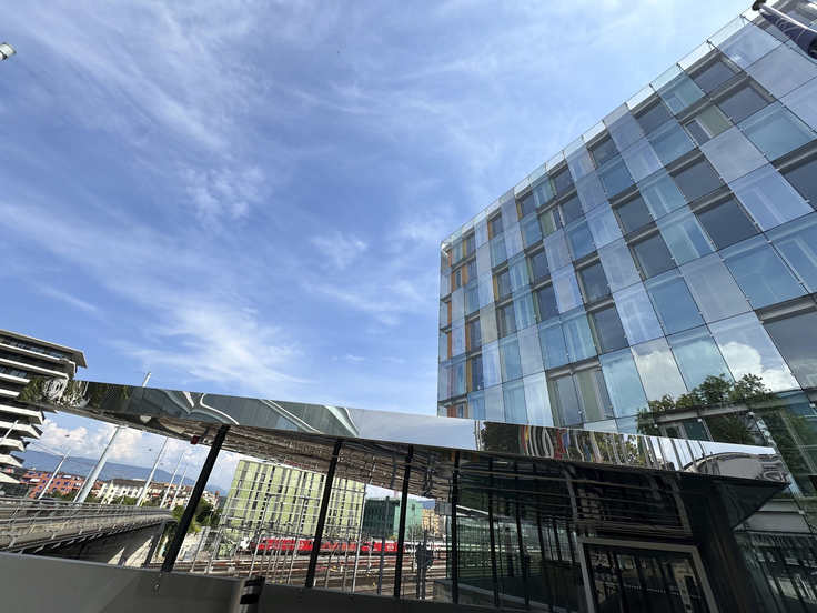  ChatGPT The photo shows a modern glass building reflecting a blue sky, a shiny canopy, and a train station with tracks and a red train in the background. The scene feels open and contemporary.