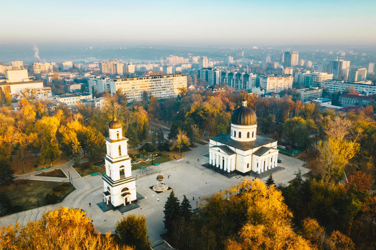 Cathedral and bell tower in a park with autumn trees, cityscape in the background.