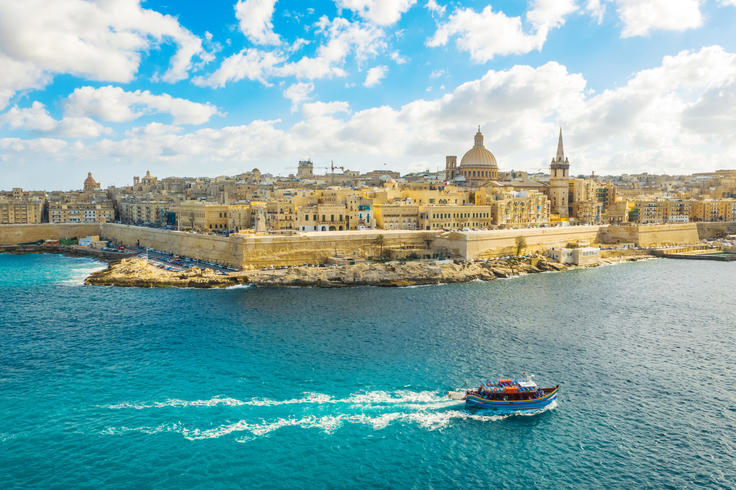 Valletta's skyline with historic buildings under a blue sky, viewed from the sea with a small boat in the foreground.
