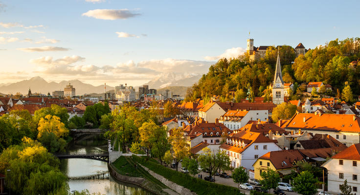 Historic city with orange rooftops, church spire, river, and lush trees under a golden evening sky.