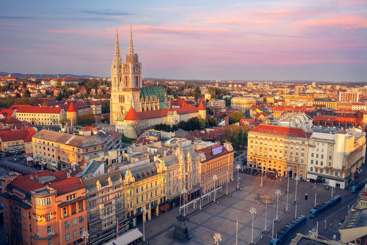 Aerial view of a city square with historic buildings and a cathedral, under a colorful sunset sky.