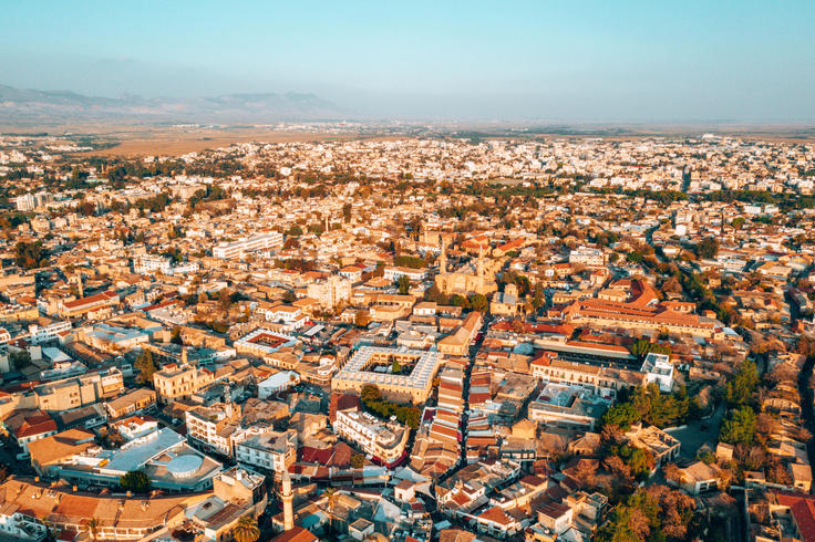 Aerial view of Nicosia, Cyprus with dense buildings and roads, set against a mountainous horizon under a clear blue sky.
