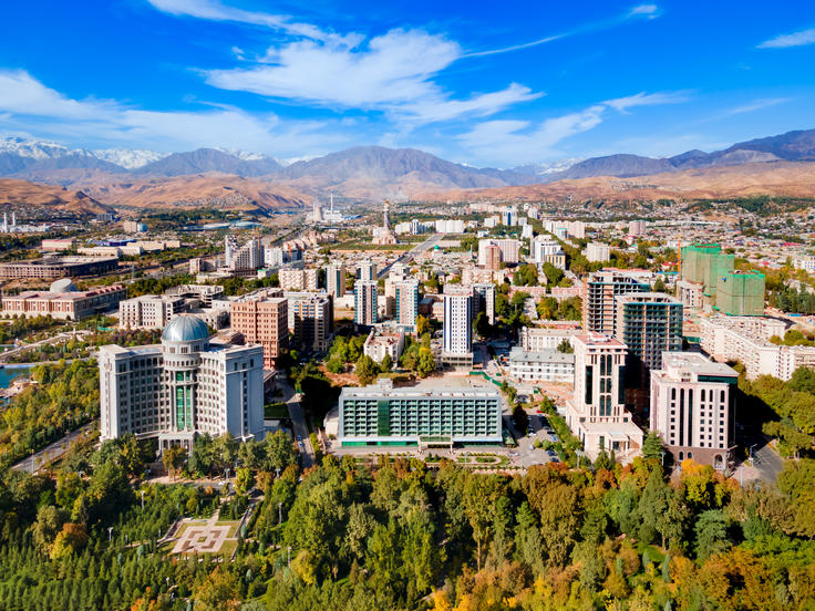 Aerial view of a cityscape with modern buildings, surrounded by green trees and mountains under a bright blue sky.
