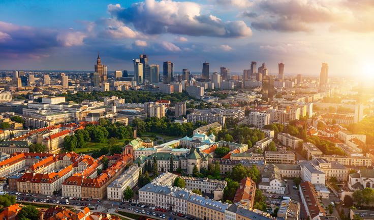 Aerial view of a cityscape at sunset, with skyscrapers, green parks, and historic buildings beneath a cloudy sky.
