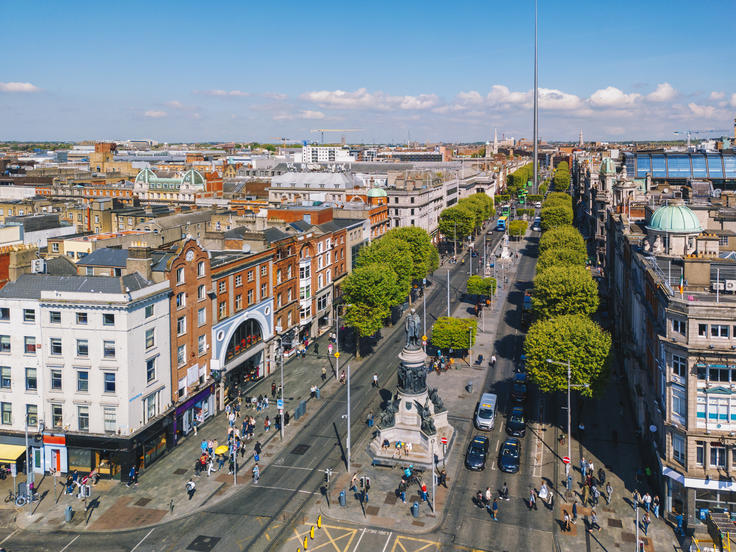 Aerial view of a busy city street lined with trees and buildings, under a clear blue sky.