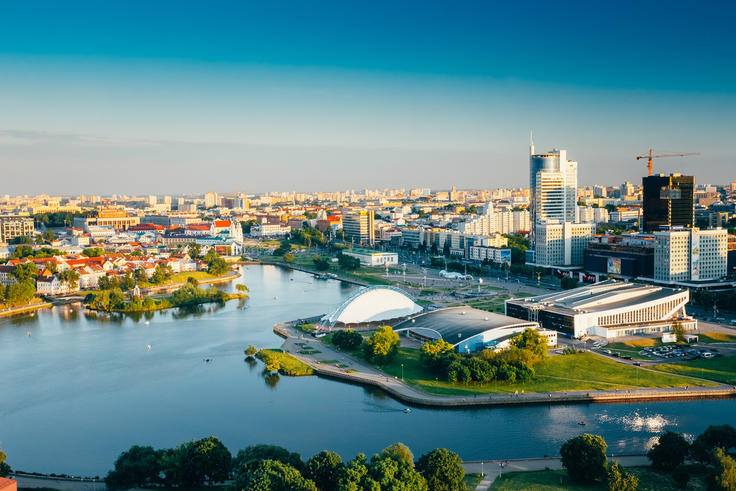 Aerial view of a city skyline with a river, modern buildings, and green spaces under a clear blue sky.