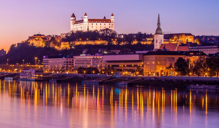 Bratislava Castle illuminated at sunset, reflecting on the Danube River, with a clear purple sky.