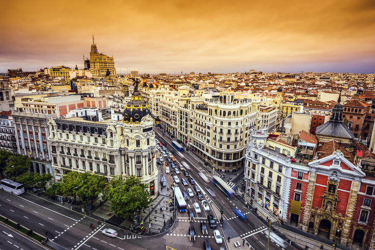 Bustling cityscape with ornate buildings under an orange sunset sky, busy street with cars and buses.