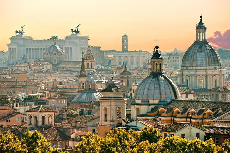 Skyline of Rome at sunset, featuring historic domes and buildings under a warm, orange sky.