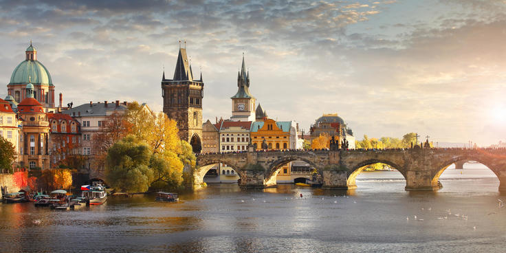 A scenic view of Charles Bridge in Prague at sunset, with historic buildings and a calm river in the foreground.