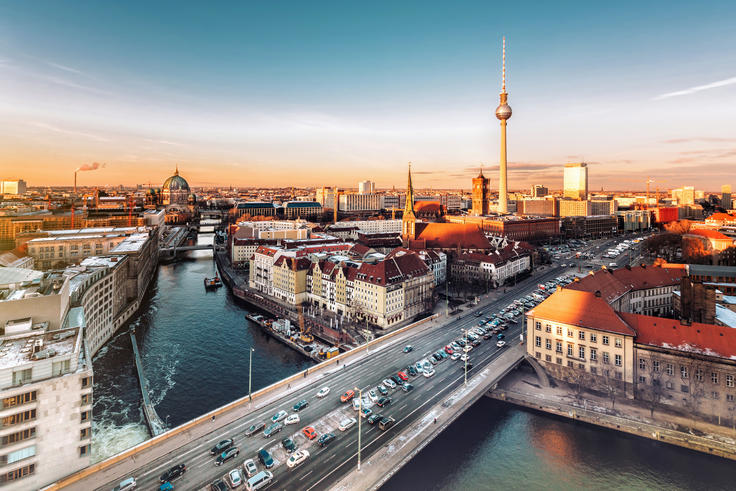Aerial view of a cityscape at sunset with a river, bridges, and a tall TV tower in the distance.