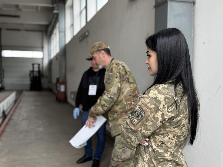 Three people in military uniforms stand in a warehouse.