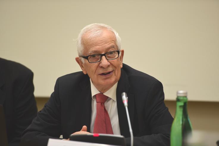 A man in a suit and tie speaks at a conference table, a microphone in front of him.