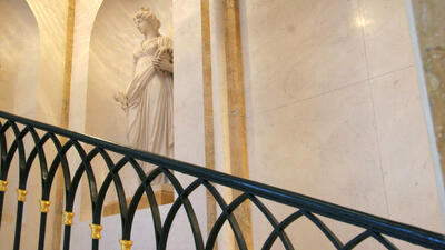 The image showcases an interior with a marble statue of a classical female figure placed in a recessed alcove. The statue is draped in flowing garments, holding an object in one hand, and wearing a crown or decorative headpiece. The background consists of polished marble walls with gold-trimmed accents. In the foreground, a decorative black wrought iron railing with gold embellishments follows the curve of a staircase. 