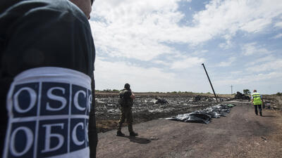 Inspectors in a field examine airplane debris, wearing uniforms with "OSCE" armbands.