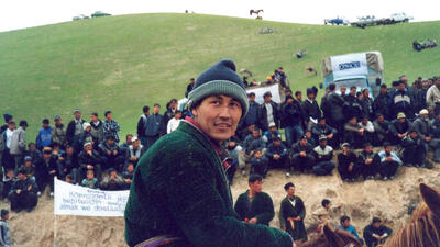 Man on horseback at an outdoor gathering with a crowd seated on a hillside and an OSCE truck in the background.