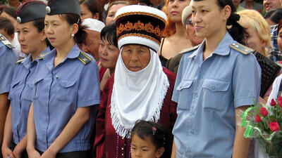 A group of women in uniform stands with a traditionally dressed older woman and a child at an outdoor event.