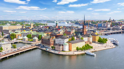 Aerial view of a vibrant cityscape by the water, with colorful buildings, a tall spire, and boats docked along the shore.