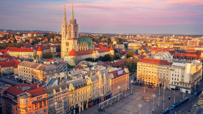 Aerial view of a city square with historic buildings and a cathedral, under a colorful sunset sky.