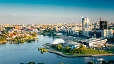 Aerial view of a city skyline with a river, modern buildings, and green spaces under a clear blue sky.