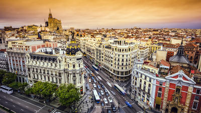 Bustling cityscape with ornate buildings under an orange sunset sky, busy street with cars and buses.