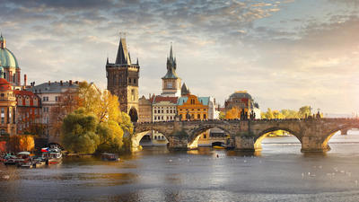 A scenic view of Charles Bridge in Prague at sunset, with historic buildings and a calm river in the foreground.