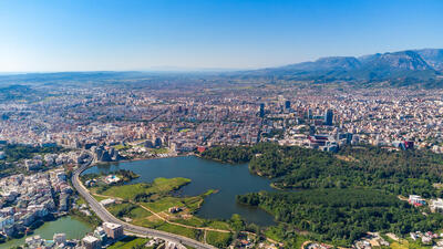 Aerial view of a city with a large green park and lake, surrounded by dense buildings, under a clear blue sky.