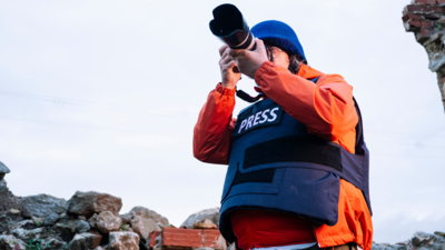 Photojournalist in an orange jacket and blue helmet, taking photos in a rocky landscape.