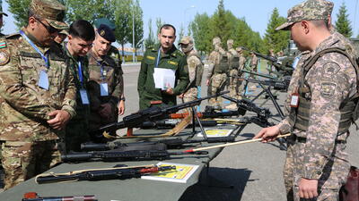 Military personnel examine displayed rifles at an outdoor exhibition, guided by an officer in camouflage uniform.