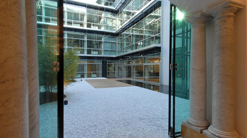 The image showcases a modern courtyard enclosed by a glass-fronted building. The perspective is from inside a classical-style room, framed by marble columns and an arched ceiling, looking out through glass doors. The courtyard features a minimalist design with a white gravel surface, a small wooden deck area, and greenery, including bamboo plants. The building’s exterior is made up of large glass panels, creating a sleek and contemporary contrast to the classical interior elements.