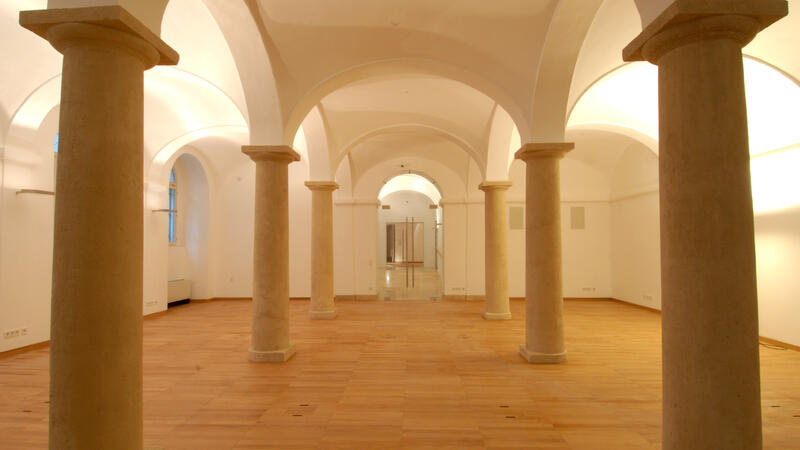 The image shows a spacious hall with classical architectural elements. The room features large, evenly spaced stone columns supporting a series of vaulted ceilings. The floor is made of warm-toned wood, contrasting with the white walls and ceilings. The lighting is soft and warm, enhancing the historical ambiance of the space.