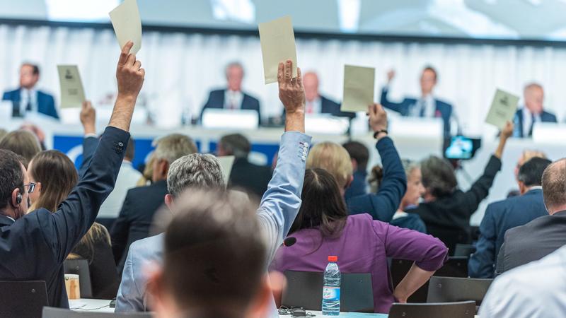 A group of Parliamentarians looking forward at a podium and raising voting cards into the air.