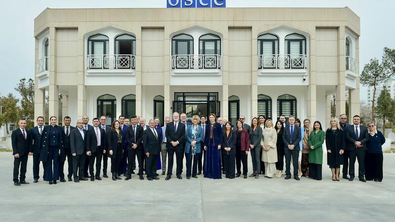 A group of people standing in front of a building with an OSCE sign, formal attire, neutral expressions.