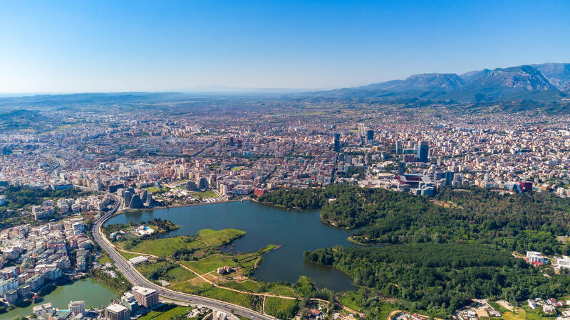 Aerial view of a city with a large green park and lake, surrounded by dense buildings, under a clear blue sky.