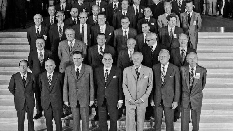 Black-and-white photograph showing the Foreign Ministers of the CSCE participating States, gathered for the first Helsinki meeting in 1973. The group is posed on a set of steps, with several rows of individuals dressed in formal suits and ties. In the background, other attendees and onlookers are visible, adding depth to the setting.