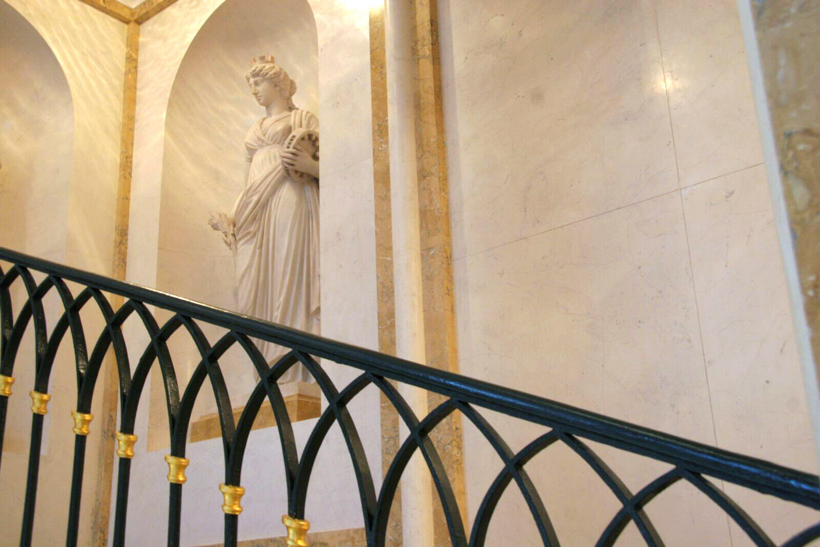 The image showcases an interior with a marble statue of a classical female figure placed in a recessed alcove. The statue is draped in flowing garments, holding an object in one hand, and wearing a crown or decorative headpiece. The background consists of polished marble walls with gold-trimmed accents. In the foreground, a decorative black wrought iron railing with gold embellishments follows the curve of a staircase. 
