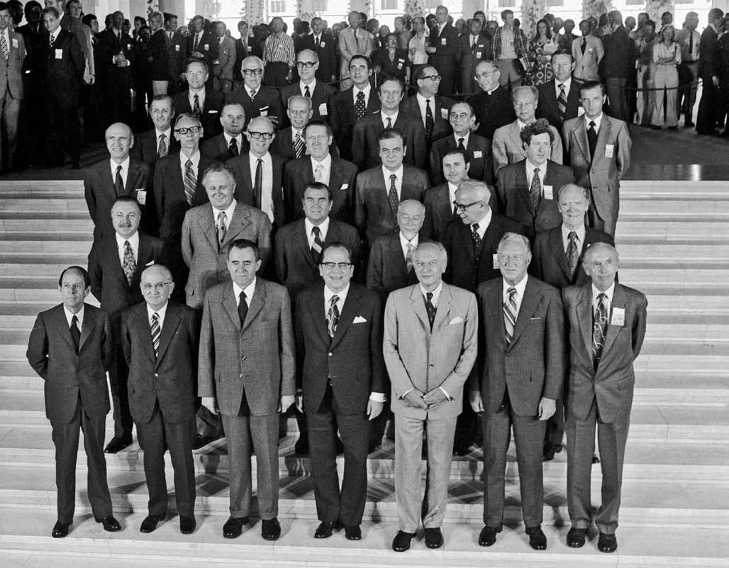 Black-and-white photograph showing the Foreign Ministers of the CSCE participating States, gathered for the first Helsinki meeting in 1973. The group is posed on a set of steps, with several rows of individuals dressed in formal suits and ties. In the background, other attendees and onlookers are visible, adding depth to the setting.